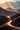 An aerial view shows a paved road winding through a desert mountain landscape. The road is prominently featured in the foreground and curves sharply to the left and then to the right. Mountain peaks are visible in the background and the image is lit by the warm glow of the morning or evening sun, with sunbeams streaming down from behind the mountains. 
