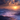 A picturesque landscape showing a sunrise over mountains. The scene is filled with rolling hills and valleys submerged in a sea of fog that appears pink from the sunlight. The sun is just peeking over the horizon, casting a warm golden glow on the clouds above the peaks.