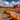A wide angle view of a pumpkin patch on a sunny day shows multiple rows of bright orange pumpkins leading toward several classic red barns in the background. Above, a blue sky is filled with fluffy white clouds. The pumpkins are set in a field of dry grasses.