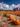 A wide angle view of a pumpkin patch on a sunny day shows multiple rows of bright orange pumpkins leading toward several classic red barns in the background. Above, a blue sky is filled with fluffy white clouds. The pumpkins are set in a field of dry grasses.