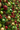 A close-up, vertically oriented image of a dense Christmas wall decoration. It features a rich tapestry of green pine needles and other foliage, interspersed with numerous ornaments, pinecones, and red berries. Prominent elements include large glittery gold spheres, deep red glittery spheres, matte and glossy green spheres, and various natural brown pinecones, creating a luxurious and festive visual texture that extends into a blurred background.