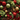 A dense, full-frame close-up of Christmas decorations. The image features a rich tapestry of deep green evergreen branches, interspersed with various sized ornaments in glittery red, shimmering gold, and matte olive green. Also visible are clusters of small red berries, brown pine cones, and faux flowers in both bright red and light green, creating a vibrant and festive holiday display.