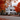 A two-story white house is centered in the frame, surrounded by trees with brilliant orange and red leaves. The house has a grey roof, black shutters, and a dark front door. The road in front of the house is covered in fallen autumn leaves.