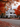 A two-story white house is centered in the frame, surrounded by trees with brilliant orange and red leaves. The house has a grey roof, black shutters, and a dark front door. The road in front of the house is covered in fallen autumn leaves.