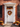 A charming front door decorated for autumn. A white door is centered in the frame, framed by red brick and dark window shutters. A vibrant fall wreath hangs on the door and garland sits above it. Orange and white pumpkins and potted mums decorate the steps leading to the door.
