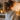 A young woman, seen from behind and her right profile, stands in a warmly lit indoor setting. She has light brown hair tied in a ponytail and is wearing a cream-colored, textured knitted sweater. Her head is turned slightly to the right, looking towards a white shelf adorned with multiple lit candles inside and outside a dark metal lantern, creating a soft, golden glow. A white radiator is partially visible at the bottom left, and a small pine branch hangs in the upper right background.