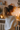 A young woman, seen from behind and her right profile, stands in a warmly lit indoor setting. She has light brown hair tied in a ponytail and is wearing a cream-colored, textured knitted sweater. Her head is turned slightly to the right, looking towards a white shelf adorned with multiple lit candles inside and outside a dark metal lantern, creating a soft, golden glow. A white radiator is partially visible at the bottom left, and a small pine branch hangs in the upper right background.