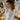 A young woman with light brown hair, wearing a cream-colored cable knit sweater, gently holds her hair up as if styling it into a ponytail. She is looking thoughtfully to the left with a soft, natural expression. The background is softly blurred but reveals warm string lights, glowing candles on a white radiator, and a wall-mounted air conditioning unit displaying '20', suggesting a cozy and festive indoor setting.