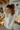 A young woman with light brown hair, wearing a cream-colored cable knit sweater, gently holds her hair up as if styling it into a ponytail. She is looking thoughtfully to the left with a soft, natural expression. The background is softly blurred but reveals warm string lights, glowing candles on a white radiator, and a wall-mounted air conditioning unit displaying '20', suggesting a cozy and festive indoor setting.