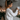 A young woman in a cream-colored chunky knit sweater adjusts a white wall-mounted smart home control panel with her right hand. Her brown hair is tied in a high ponytail, and she is shown in profile against a textured grey wall. The background features warm, glowing candles, green pine garland, and decorative lanterns, creating a cozy and inviting atmosphere beside a white radiator.