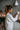 A young woman in a cream-colored chunky knit sweater adjusts a white wall-mounted smart home control panel with her right hand. Her brown hair is tied in a high ponytail, and she is shown in profile against a textured grey wall. The background features warm, glowing candles, green pine garland, and decorative lanterns, creating a cozy and inviting atmosphere beside a white radiator.