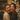 A close-up portrait of two young women hugging and laughing with their eyes closed, beaming with genuine happiness. One woman wears a cream knit sweater, the other a dark red speckled knit sweater. In the softly blurred background, a warm, glowing Christmas tree with bokeh lights creates a festive and cozy atmosphere.