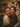A close-up portrait of two young women hugging and laughing with their eyes closed, beaming with genuine happiness. One woman wears a cream knit sweater, the other a dark red speckled knit sweater. In the softly blurred background, a warm, glowing Christmas tree with bokeh lights creates a festive and cozy atmosphere.