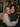 A close-up vertical shot features two young women embracing and laughing heartily, their faces beaming with joy. The woman on the left has blonde, wavy hair and wears a light-colored, textured knit sweater, while the woman on the right has brown hair, visible freckles, and wears a dark grey knit sweater and a dark red beanie. Behind them, a Christmas tree with blurred, warm golden string lights creates a festive bokeh background, adding to the cozy and celebratory mood.