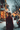A vertical shot from behind a woman with light brown hair styled in a bun, wearing a dark red, possibly velvet, winter coat. She is walking forward through a snowy environment. Numerous out-of-focus warm golden and orange string lights illuminate the blurry urban background, creating a festive bokeh effect, while white snowflakes fall gently around her. The ground is covered in snow, suggesting a serene winter evening in a decorated city street or market.