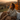 A woman with long brown hair, wrapped in a brown knitted blanket and a cream-colored sweater, is sitting on a wicker couch. She is holding a white cup and looking off to the left with a happy smile. Behind her, the sun is setting, creating a golden glow over the distant hills and foliage.