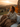 A woman with long brown hair, wrapped in a brown knitted blanket and a cream-colored sweater, is sitting on a wicker couch. She is holding a white cup and looking off to the left with a happy smile. Behind her, the sun is setting, creating a golden glow over the distant hills and foliage.