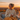 A woman is sitting in a wicker chair outside, wrapped in a large yellow blanket and holding a mug in her hands. She is smiling and looking off to the side. The background features a sunset with orange and yellow hues, creating a warm and inviting atmosphere.