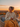 A woman is sitting in a wicker chair outside, wrapped in a large yellow blanket and holding a mug in her hands. She is smiling and looking off to the side. The background features a sunset with orange and yellow hues, creating a warm and inviting atmosphere.