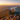 A woman with medium-length brown hair stands on a rocky outcrop at the peak of a mountain, looking out at a vast scenic view. She is wearing a bright yellow jacket, black leggings, and hiking boots. The landscape includes rolling mountains covered in trees, a large lake in the distance, and a vibrant sunset casting golden light across the entire scene.