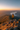 A woman with medium-length brown hair stands on a rocky outcrop at the peak of a mountain, looking out at a vast scenic view. She is wearing a bright yellow jacket, black leggings, and hiking boots. The landscape includes rolling mountains covered in trees, a large lake in the distance, and a vibrant sunset casting golden light across the entire scene.