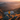 A woman stands on a rocky mountain ridge looking at a panoramic view. The landscape includes mountains layered into the distance, with a calm lake nestled in the valley below. A warm sunrise creates golden light bathing the grassy hillside and the woman's yellow jacket.