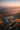 A woman stands on a rocky mountain ridge looking at a panoramic view. The landscape includes mountains layered into the distance, with a calm lake nestled in the valley below. A warm sunrise creates golden light bathing the grassy hillside and the woman's yellow jacket.