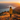A woman stands on a mountain top, facing away from the viewer, and looks out at a long lake and mountains in the distance. She is wearing a bright yellow jacket, and the scene is bathed in the warm glow of sunset, with the sun just visible on the right side. The hillside is covered in golden grass and small patches of red plants.