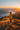 A woman stands on a mountain top, facing away from the viewer, and looks out at a long lake and mountains in the distance. She is wearing a bright yellow jacket, and the scene is bathed in the warm glow of sunset, with the sun just visible on the right side. The hillside is covered in golden grass and small patches of red plants.