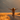 A woman with curly brown hair rides a bicycle away from the viewer on a narrow asphalt road. Her arms are spread wide, and she wears a floral shirt and denim shorts. To each side of the road is dry golden grass, and in the distance are softly blurred mountain ranges under a warm, golden sky.