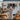 A smiling woman stands in front of a white, rustic pumpkin stand, decorated with autumn elements. She wears a blue shirt, striped apron, and brown boots and is surrounded by hay bales and pumpkins. The scene evokes a feeling of warmth, harvest, and small-town charm.