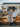 A smiling woman stands in front of a white, rustic pumpkin stand, decorated with autumn elements. She wears a blue shirt, striped apron, and brown boots and is surrounded by hay bales and pumpkins. The scene evokes a feeling of warmth, harvest, and small-town charm.