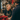 A blonde woman with a bright smile, wearing a dark blue sweater and a striped apron, stands behind a wooden counter, looking towards a customer whose back is visible. The festive shop is adorned with Christmas garlands, a large red bow, and a red poinsettia plant in the foreground, creating a warm and welcoming holiday shopping scene with soft, ambient lighting.
