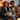 A smiling African American couple sit closely together on a gray couch with their black and white border collie dog nestled between them. The woman holds a TV remote. Warm, soft light illuminates the scene, with the glow of candles and decorative pumpkins in the foreground adding a cozy ambiance. The couple are both looking off to the right of the frame with smiles on their faces.