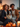 A smiling African American couple sit closely together on a gray couch with their black and white border collie dog nestled between them. The woman holds a TV remote. Warm, soft light illuminates the scene, with the glow of candles and decorative pumpkins in the foreground adding a cozy ambiance. The couple are both looking off to the right of the frame with smiles on their faces.
