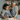 An African American grandmother and her biracial granddaughter are happily engaged in baking activities in a warm, inviting kitchen. The granddaughter uses a wooden rolling pin to flatten cookie dough on a floured surface, while the grandmother stands beside her, smiling affectionately. Both wear dark gray aprons with white snowflake patterns, set against a background of shelves softly lit by candles, suggesting a festive holiday atmosphere.