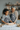An African American grandmother and her biracial granddaughter are happily engaged in baking activities in a warm, inviting kitchen. The granddaughter uses a wooden rolling pin to flatten cookie dough on a floured surface, while the grandmother stands beside her, smiling affectionately. Both wear dark gray aprons with white snowflake patterns, set against a background of shelves softly lit by candles, suggesting a festive holiday atmosphere.
