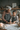 An older Black woman with gray curly hair and glasses smiles down at a young mixed-race girl as they stand side-by-side in a cozy kitchen. Both are wearing black aprons with white snowflakes. The girl is actively using a wooden rolling pin to flatten dough on a floured wooden table, where several star-shaped cookie cutouts are already visible. A soft, warm glow from a blurred lit candle is visible in the foreground to the left.
