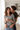 A smiling woman in a black and white striped shirt is holding a white mug while being hugged by a man from behind in a bright, modern kitchen. The man is kissing the woman's cheek, and both appear happy and content. The background includes white cabinets and a window with sheer curtains allowing natural light to flood the scene.