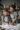 A smiling mother and her young daughter, both wearing matching grey sweaters and snowflake aprons, stand together in a rustic kitchen, hands covered in flour as they knead dough on a wooden table. Soft, warm lighting illuminates their joyful expressions and the cozy, festive background adorned with candles and string lights, capturing a moment of loving family connection during holiday baking.