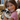 A young girl with short brown hair and orange painted cheeks smiles while holding a black and white spotted rabbit. She's wearing a pink shirt, and the background suggests a farm or petting zoo environment, including hay bales and other animals. The scene conveys a feeling of joy and connection between a child and an animal.