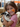 A young girl with short brown hair and orange painted cheeks smiles while holding a black and white spotted rabbit. She's wearing a pink shirt, and the background suggests a farm or petting zoo environment, including hay bales and other animals. The scene conveys a feeling of joy and connection between a child and an animal.