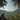 A brown monkey sits in profile on a dark, rocky outcrop in the lower-left foreground, looking to the right. The monkey overlooks a vast, dense tropical jungle valley filled with countless palm trees, stretching towards distant green mountains under a clear blue sky with scattered white clouds. Tall palm tree trunks frame the left and right edges of the image, with their fronds partially obscuring the upper corners, creating a natural frame for the panoramic scene.
