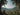 A brown monkey sits in profile on a dark, rocky outcrop in the lower-left foreground, looking to the right. The monkey overlooks a vast, dense tropical jungle valley filled with countless palm trees, stretching towards distant green mountains under a clear blue sky with scattered white clouds. Tall palm tree trunks frame the left and right edges of the image, with their fronds partially obscuring the upper corners, creating a natural frame for the panoramic scene.
