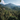 A wide, high-angle view of a dense tropical mountain range under a blue sky with some white clouds. The entire scene is covered in lush green foliage, featuring a foreground framed by large palm fronds and a midground filled with countless coconut palm trees. Distant mountains reveal rocky sections amidst the dense tree cover, conveying a wild and natural, untouched environment.