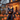 An outdoor scene featuring Halloween decorations. A large, inflatable black cat sits prominently in the foreground, surrounded by many lit jack-o'-lanterns. In the background, a house is visible, decorated with warm string lights, casting a festive glow on the lawn.