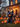 An outdoor scene featuring Halloween decorations. A large, inflatable black cat sits prominently in the foreground, surrounded by many lit jack-o'-lanterns. In the background, a house is visible, decorated with warm string lights, casting a festive glow on the lawn.