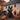 A large, black inflatable cat sits in the front yard of a white farmhouse. Carved pumpkins with lit faces are scattered around the cat and the yard. The farmhouse is decorated with pumpkins and autumn foliage, creating a festive Halloween scene.