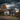 The image shows an old, two-story farmhouse with weathered white siding, set against a backdrop of dark, stormy clouds. The house is surrounded by autumn foliage with hues of orange and yellow. Several small pumpkins are seen next to the house. The windows are broken and the house appears to be in a state of disrepair, adding to the eerie and abandoned feel of the scene.