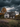 The image shows an old, two-story farmhouse with weathered white siding, set against a backdrop of dark, stormy clouds. The house is surrounded by autumn foliage with hues of orange and yellow. Several small pumpkins are seen next to the house. The windows are broken and the house appears to be in a state of disrepair, adding to the eerie and abandoned feel of the scene.